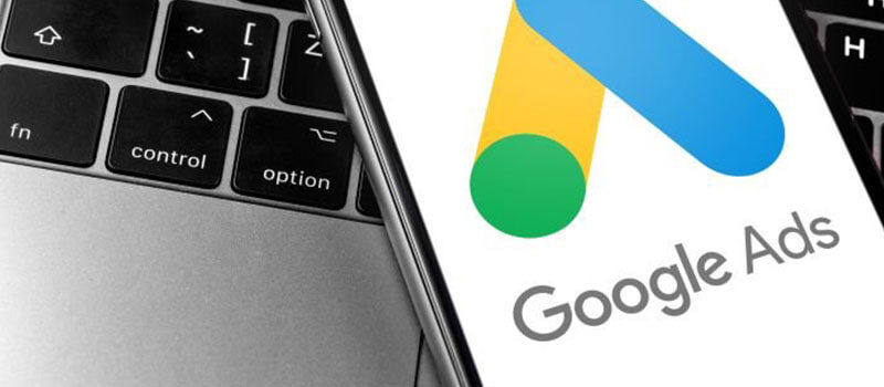 Mobile phone displaying the Google Ads logo with a laptop in the background